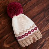 The burgunday, natural, and fawn colour fairisle hand knitted hat lying flat on a dark wooden table