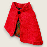 Side view of red handknitted merino baby cape with black button and tartan lining.