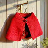 Red merino baby capelet with tartan lining, displayed on a hanger.