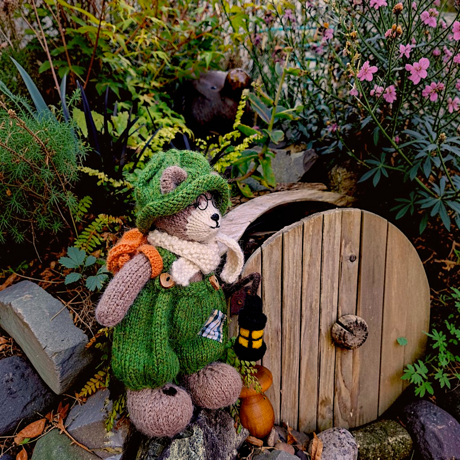 Pip Elder, the first Pebblewood Resident, a hand-knitted collectible bear with glasses and a felted lantern, beside the handmade hobbit door in Pebblewood.