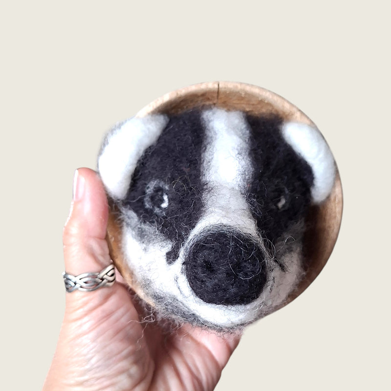 Hand needle-felted black and white badger face mounted in a small wooden bowl, shown in hand to demonstrate its scale.