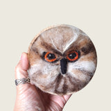 Hand needle-felted brown owl face with orange eyes mounted in a small wooden bowl, shown in hand to demonstrate its scale.