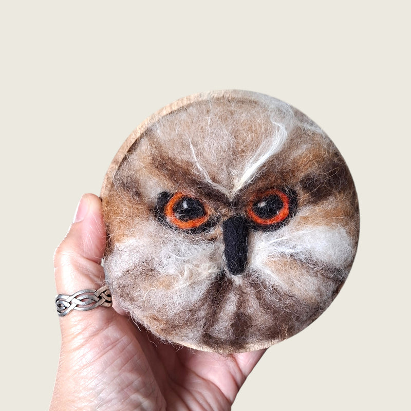 Hand needle-felted brown owl face with orange eyes mounted in a small wooden bowl, shown in hand to demonstrate its scale.