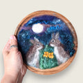 Hand needle-felted rabbits together under a moonlit sky, mounted in a wooden bowl and shown in hand to demonstrate its scale.