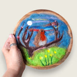 Hand needle-felted robin perched on a branch in a colourful landscape, mounted in a wooden bowl and shown in hand to demonstrate its scale.