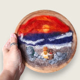 Hand needle-felted seaside sunset scene with two figures sitting on a rocky shore, mounted in a wooden bowl and shown in hand to demonstrate its scale.