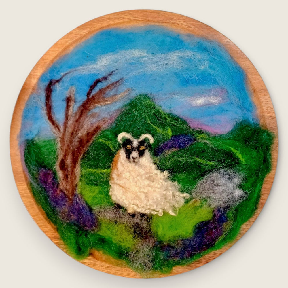 Needle felted wool picture of a sheep standing on a hillside beneath a blue sky, mounted on a round wooden plaque and handmade in Ireland.
