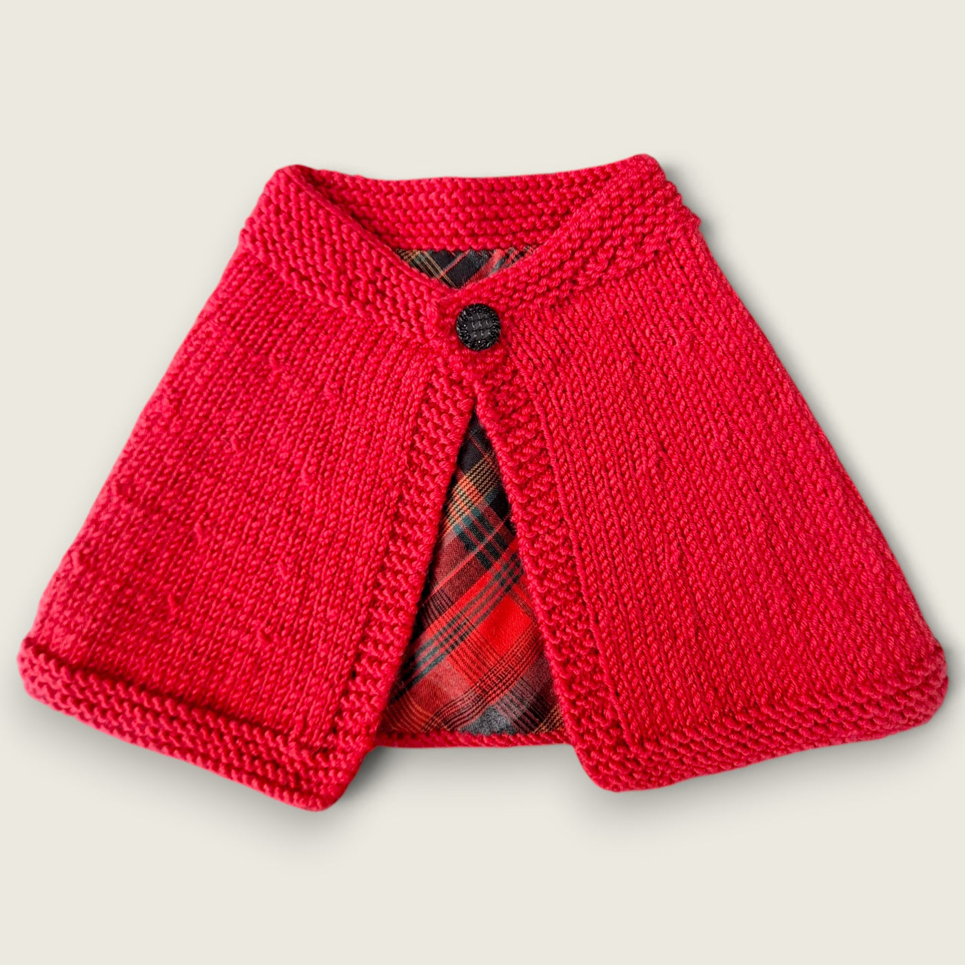Red handknitted cape with black button fastening and tartan lining.