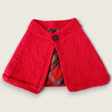 Red handknitted cape with black button fastening and tartan lining.
